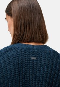 Frau mit glattem braunem Haar, die einen dunkelblauen grob gestrickten Pullover trägt, von hinten sichtbar mit einem kleinen metallischen Markenschild am Ausschnitt.