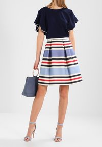 Mörkblå blus med volangärmar i kombination med en randig knä-lång kjol i blått, rött och krämfärgat. Accessoriserad med en blå handväska.