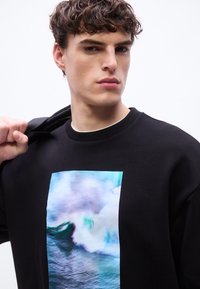 Νέος άνδρας με σγουρά μαλλιά που φοράει μαύρο φούτερ με ένα μπλε γραφικό κύμα ωκεανού, κρατώντας ένα μπουφάν στον ώμο του.