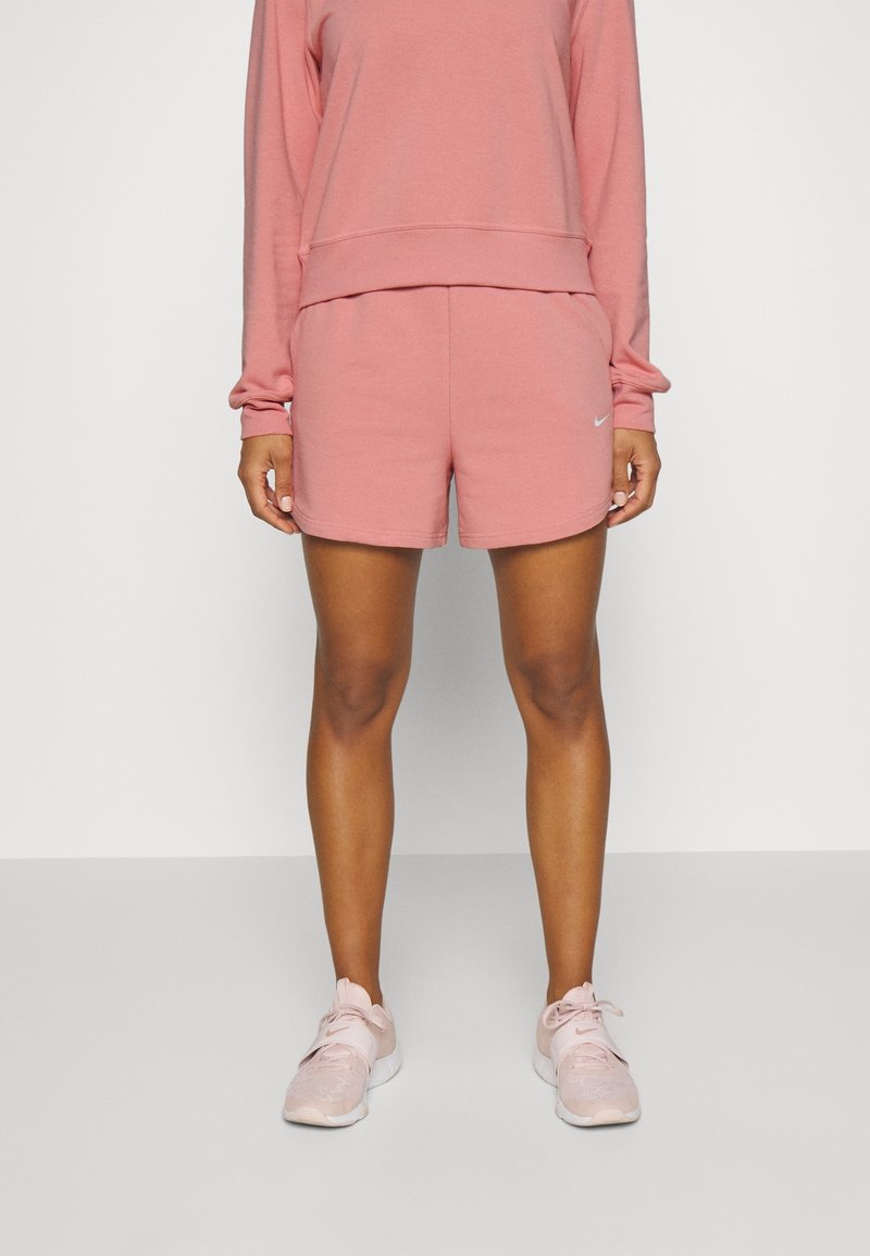 Personne portant un haut à manches longues rose, un short rose assorti avec le logo Nike, et des baskets rose clair, debout devant un fond blanc uni.