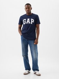 Navy t-shirt with white "GAP" logo, short sleeves, paired with loose-fit distressed blue jeans and grey sneakers with black stripes.