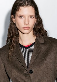 Brown wool-blend blazer with a single button, paired with a striped black and white top featuring a red accent, smooth texture.