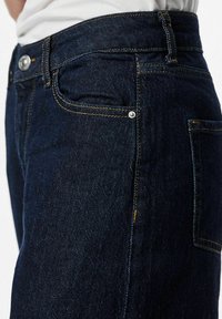 Tumši zilo džinsu bikses ar dzelteno diegu, redzamām kabatām un metāla pogu jostā. Tuvplāns par auduma faktūru.