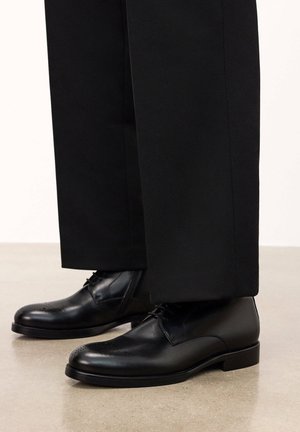 Schwarze Lederschuhe mit glatter Oberfläche, abgerundeter Spitze, dezenten Perforationen und niedrigem, gestapeltem Absatz. Getragen mit schwarzen Hosen.