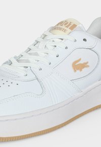 White Lacoste sneaker with beige crocodile logo, white laces, and light brown sole, showing detailed stitching and perforations.
