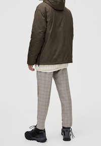Oliven pufferjakke med hætte, off-white strikunderlag og gråternet bukser. Sorte robuste sko fuldender outfittet.