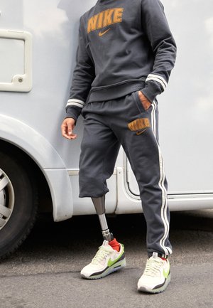 Persoon die een donkergrijs Nike trainingspak met witte strepen draagt, staat naast een voertuig, toont een prothetisch been en witte Nike sneakers met groene accenten.