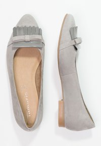 Un par de mocasines de ante gris claro con tacones bajos y correas decorativas de flecos grises brillantes sobre un fondo blanco.
