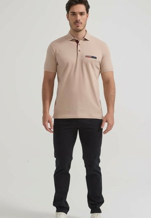 Homme debout portant un polo beige à manches courtes avec une tache sombre sur la poche poitrine et un pantalon noir sur un fond uni.