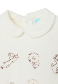 Crème-kleurig kledingstuk met een zachte, pluche kraag. Bevat bruine geborduurde cartoonfiguren in verschillende poses op de voorkant.