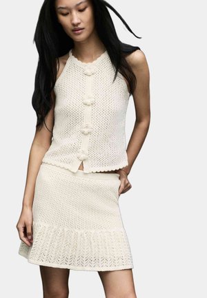 Donna che indossa un top in maglia crema senza maniche con bottoni a forma di fiore e una gonna coordinata in maglia con orlo strutturato, posando con una mano in tasca.