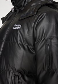 Czarna pikowana kurtka o wyglądzie skóry z przednim zamkiem błyskawicznym, klapką na zatrzaski, kieszenią na rękawie z zamkiem oraz białym haftowanym napisem "NIGHT ADDICT" na piersi.