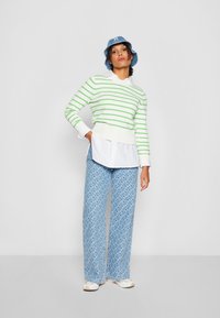 Grün gestreifter Pullover mit weißem Kragen, über einem weißen Hemd getragen. Weite, blaue Hosen mit Muster, ergänzt durch einen blauen Bucket Hat.
