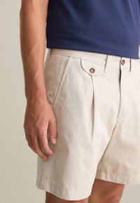 Pantalones cortos plisados beige con bolsillos laterales, con una textura suave y botones decorativos. Llevados con una camisa de manga corta azul marino.