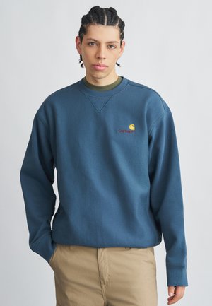 Blaues Baumwoll-Sweatshirt mit geripptem Rundhalsausschnitt, versehen mit einem kleinen gestickten Logo und legerer Passform. Kombiniert mit beigen Hosen.