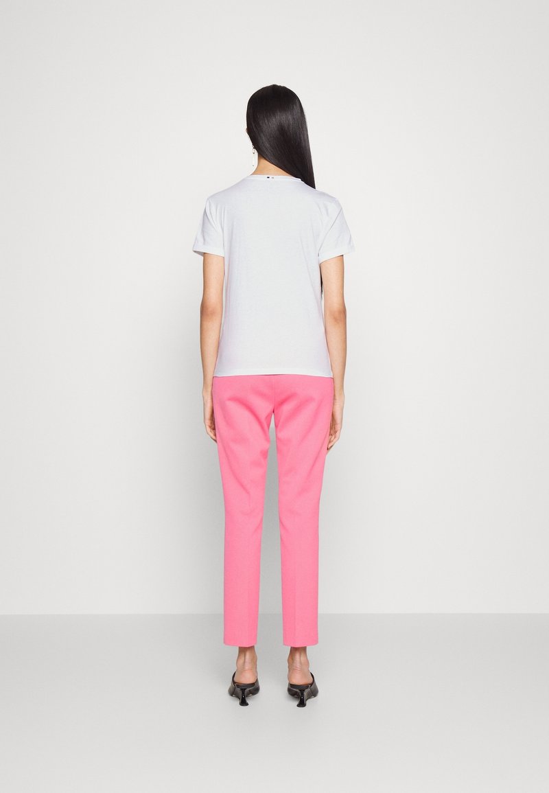 Femme debout, dos à la caméra, portant une chemise blanche à manches courtes, un pantalon rose et des chaussures à talons noires, sur un fond uni.