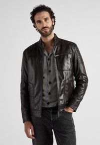 Svart läderjacka med krage, framdragkedja och texturerade axlar, kombinerad med en randig skjorta och mörka jeans.