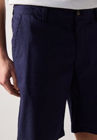 Sötétkék, könnyű anyagból készült rövidnadrág, lapos elülső résszel, oldalsó zsebekkel és gombos záródással. Simázott felület, letisztult vonalakkal.