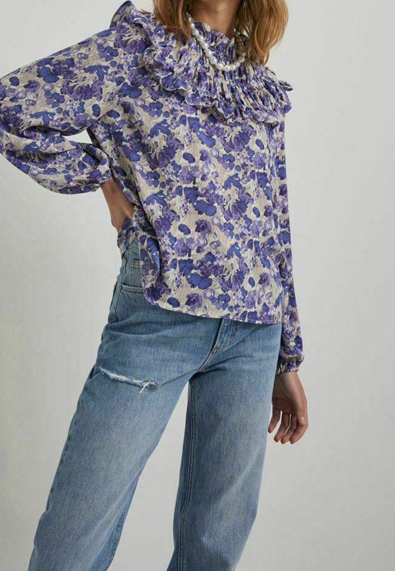 Femme portant un chemisier ample à volants à fleurs violet, associé à un jean taille haute bleu clair avec une petite déchirure sur la cuisse.