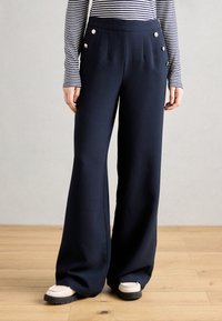 Pantalons larges bleu marine en tissu lisse, ornés de boutons dorés et de plis à l'avant. Associés à des chaussures légères.