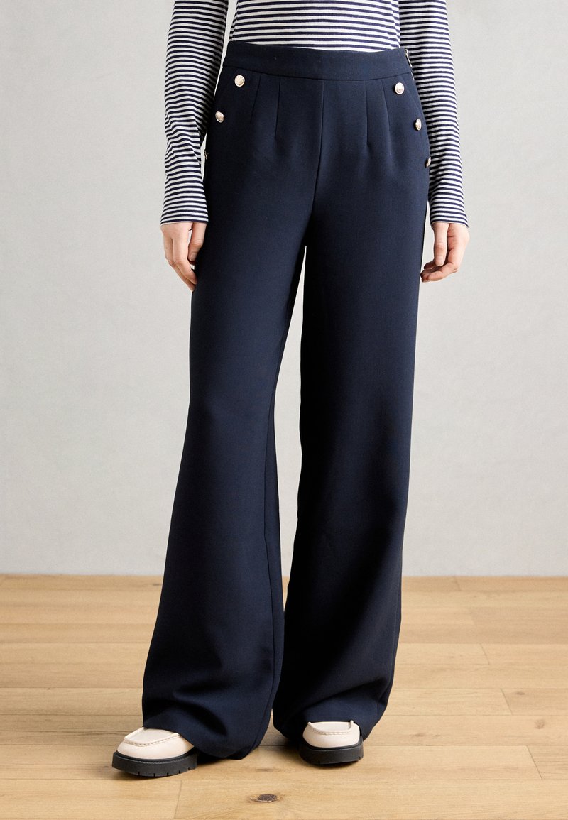 Pantalons larges bleu marine en tissu lisse, ornés de boutons dorés et de plis à l'avant. Associés à des chaussures légères.