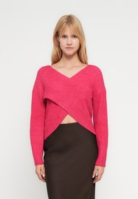 Krótki różowy prążkowany sweter z dekoltem w serek i kopertowym wzorem. Ma długie rękawy i jest zestawiony z brązową ołówkową spódnicą.