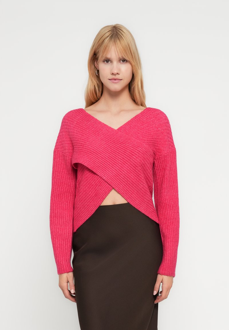 Krótki różowy prążkowany sweter z dekoltem w serek i kopertowym wzorem. Ma długie rękawy i jest zestawiony z brązową ołówkową spódnicą.