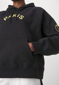 Sweat à capuche noir avec un logo jaune "PARIS" sur la poitrine et une poche à l'avant. Poignets et ourlet côtelés. Texture de tissu douce.
