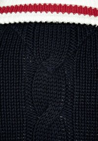 Mörkblå stickad tröja med kabelmönster, med en vit och röd ribbad krage. Strukturerad tyg med distinkta sömmar och design.