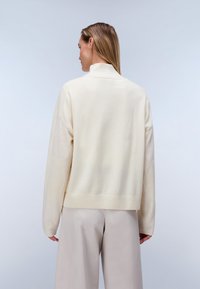 Kvinde med langt, glat hår iført en kremfarvet rullekravesweater og lysebeige bukser med vide ben, vist bagfra.