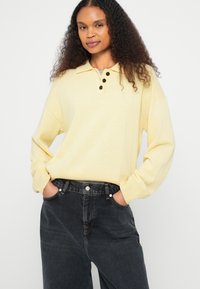 Kollane pikakäteline polo-särk kraega ja kolme musta nupuga, kombineeritud kõrge vöökohaga tumedate teksadega. Sile tekstuur.