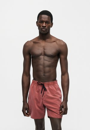 Muž s svalnatou postavou, ktorý má na sebe hrdzavé plavkové šortky s čiernym šnúrkom, stojí na jednoduchom svetlosivom pozadí.