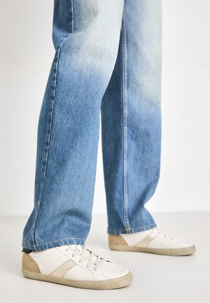 Jambes vêtues d'un jean droit bleu clair délavé, associées à des baskets blanches avec du daim beige et un accent doré.