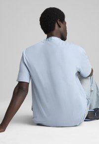 Lyseblå kortærmet t-shirt lavet af blødt stof, med en rund halsudskæring og et lille grønt mærke med teksten "Motorsport" bagpå.
