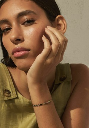 Frau mit glatter Haut, die ihr Kinn auf die Hand stützt, trägt ein grünes ärmelloses Oberteil mit braunen Knöpfen und ein Perlenarmband am Handgelenk.