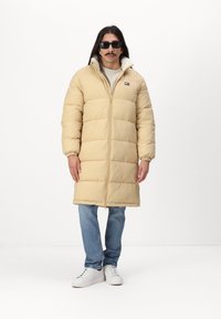 Beige pufferjack met een ritssluiting aan de voorkant, voorzien van horizontale stiksels en een hoge kraag. Draagt over een lichtgekleurde trui en blauwe jeans.