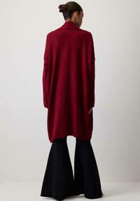 Hosszú piros kardigán sállal, puha anyagból készült. Laza szabású, oldalsó hasítékokkal. Fekete harangszárú nadrág fölött viselve.