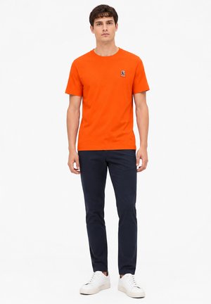 Jeune homme debout portant un t-shirt orange à manches courtes, un pantalon bleu marine foncé et des baskets blanches sur un fond blanc uni.
