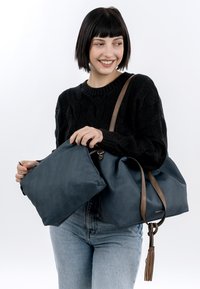 Jeune femme aux cheveux courts noirs portant un pull noir et un jean, tenant un grand sac bleu avec des sangles marron et une pochette assortie.