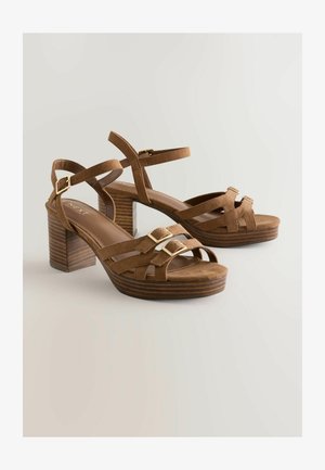 Brūni zamšādas augstpapēžu sandales ar koka bloka papēdi un krustveida siksnām, kurām priekšpusē ir divi zelta krāsas sprādzes akcenti.