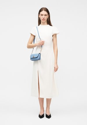 Γυναίκα με λευκό μίντι φόρεμα με κοντά μανίκια και άνοιγμα μπροστά, κρατώντας μπλε τσάντα ώμου και φοράει μαύρα μυτερά παπούτσια, στέκεται μπροστά σε λευκό φόντο.