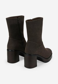 Botas de ante marrón oscuro hasta la mitad de la pantorrilla, con tacones cuadrados y cremalleras laterales, mostradas desde la parte de atrás y ligeramente en ángulo sobre un fondo blanco.