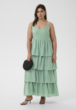 Frau trägt ein ärmelloses, mehrlagiges Maxikleid in Minzgrün, hält eine runde schwarze, geflochtene Clutch und trägt schwarze, geschnürte Sandalen mit Absatz vor schlichtem Hintergrund.