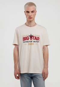 Bežinė medvilninė marškinė su spalvinga grafika, kurioje užrašas "BIG STAR" raudonai ir mėlynai, apykakle ir trumpomis rankovėmis. Šviesiai mėlynos džinsinės kelnės.