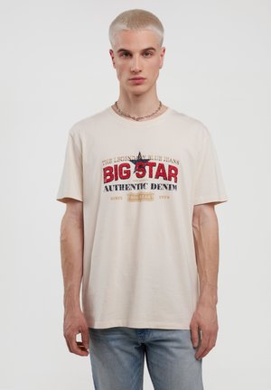 Beige bomulds T-shirt med et farverigt grafisk print med "BIG STAR" i rød og blå, rund hals, og korte ærmer. En par lys blå jeans.
