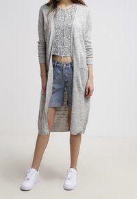 Ljusgrå cardigan med en lång, öppen design, i kombination med en fläckig, kort topp och fransade denim-shorts. Vita sneakers fulländar looken.