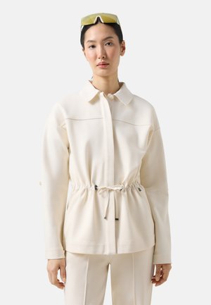 Kobieta ubrana w kremową kurtkę z sznurkiem w talii i dopasowane spodnie, z przezroczystymi żółtymi okularami przeciwsłonecznymi na głowie.