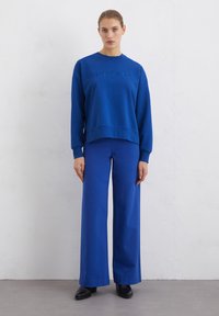 Blue sweatshirt with embossed logo and crew neckline, paired with matching wide-leg trousers and black ankle boots. Smooth fabric texture.
