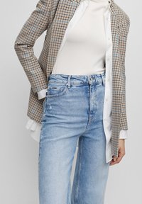 Vaaleansiniset farkut, joissa on löysä malli ja kulutettu yksityiskohta, yhdistettynä beigekuvioiseen bleiseriin ja valkoiseen yläosaan sen alla.