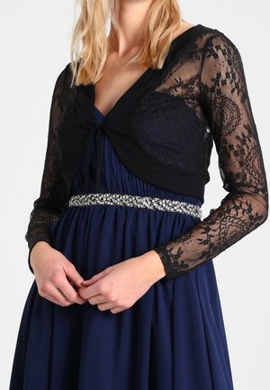 Femme portant une robe plissée bleu marine avec une ceinture ornée de bijoux et un boléro en dentelle noire à manches longues.